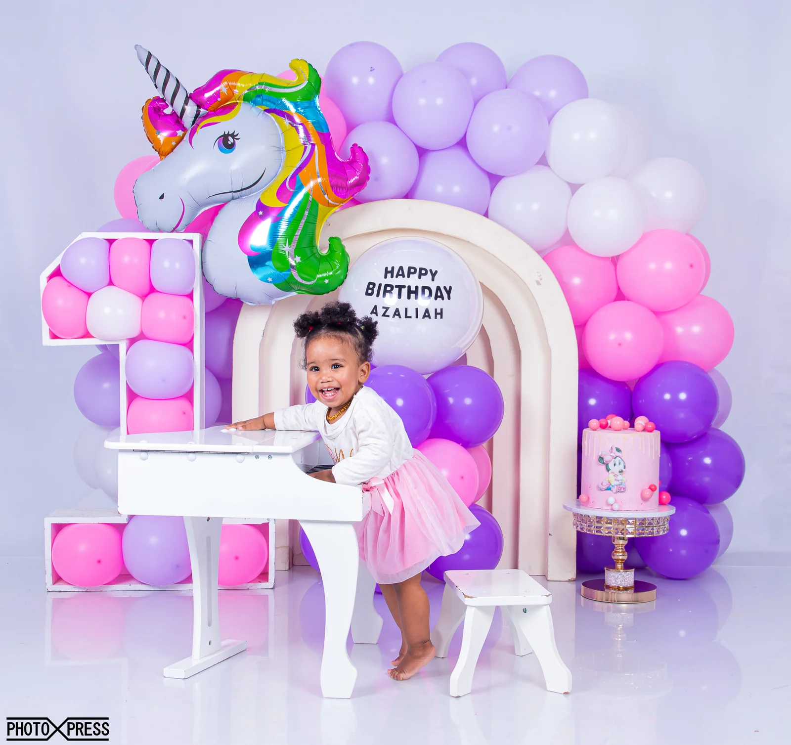 A toddler laughing at her first-birthday cake smash session, surrounded by a purple and pink balloon arch